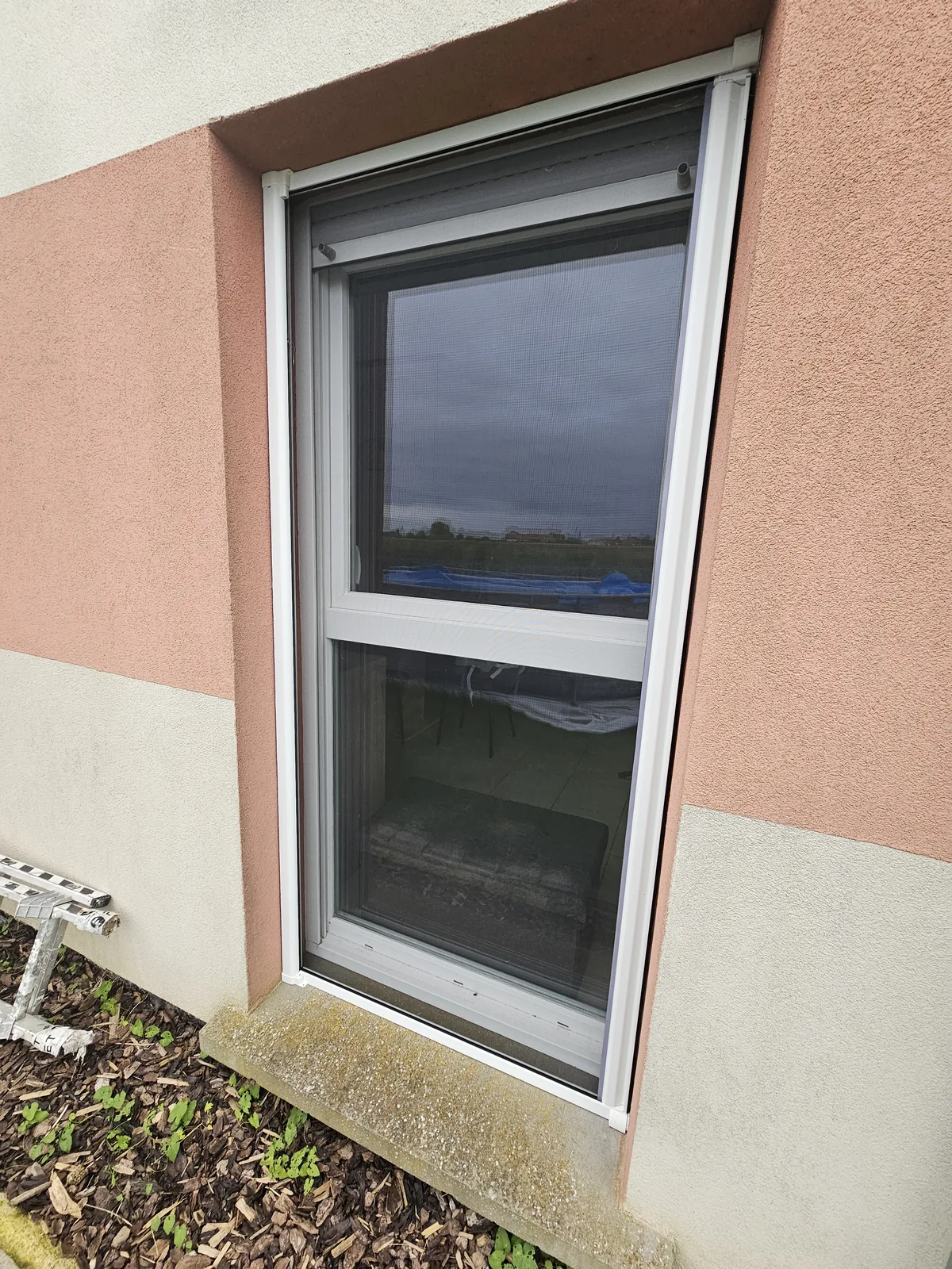 Moustiquaire coulissante blanche posée devant une porte-fenêtre, façade crépie rose et beige, rail bas sur l’appui.