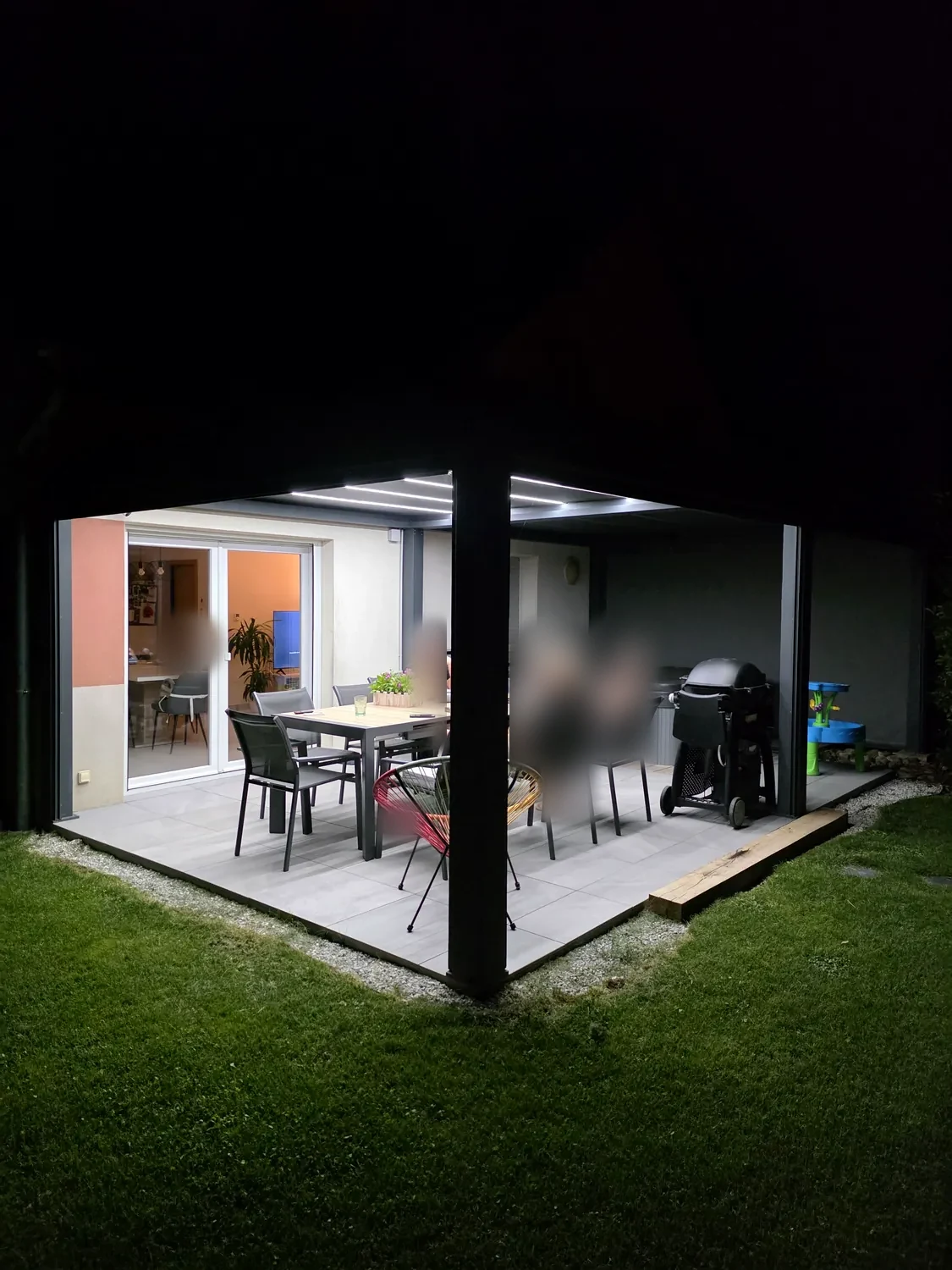 Pergola bioclimatique anthracite de nuit, lames fermées et LED allumées au-dessus d’une table dressée, terrasse dallée bordée de pelouse.