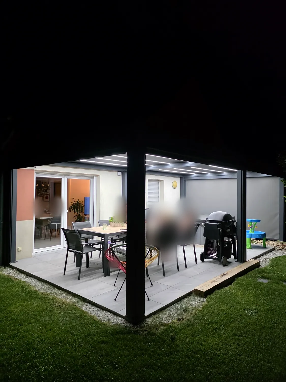 Terrasse dressée sous une pergola bioclimatique éclairée par les LED intégrées aux lames, sol dallé, baie vitrée ouverte sur la maison.