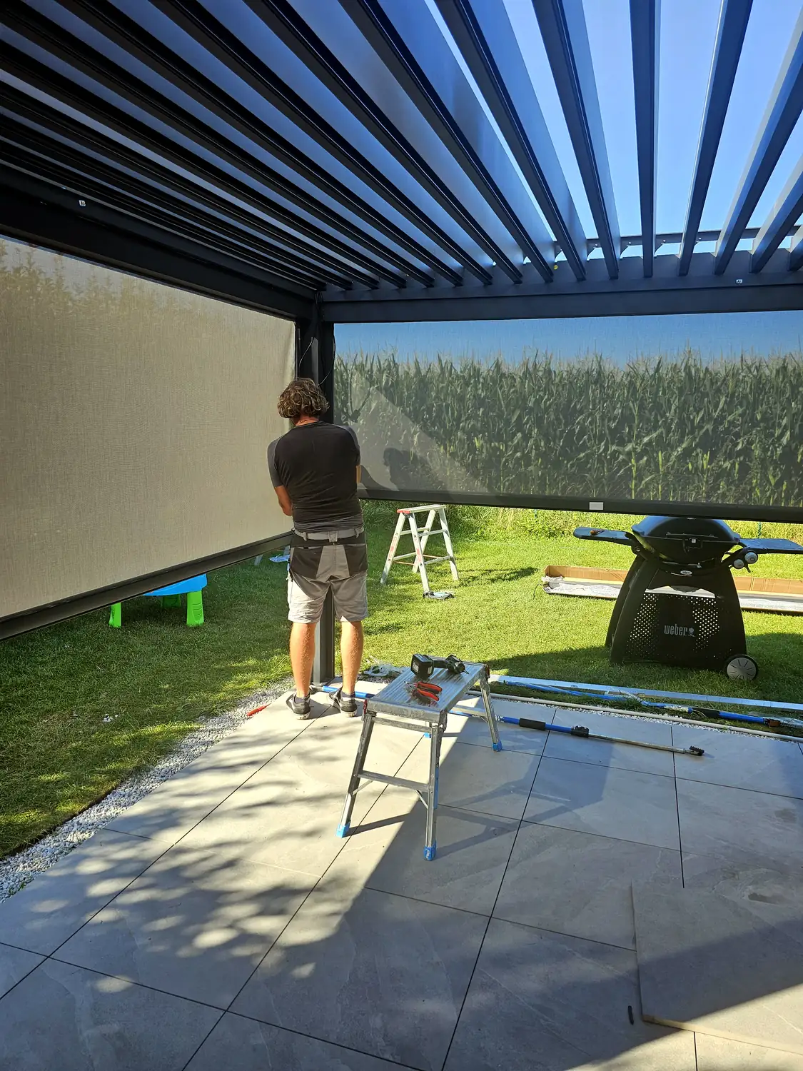 Poseur de dos réglant un store latéral sous une pergola noire, lames ouvertes sur le ciel, escabeaux et profilés au sol.