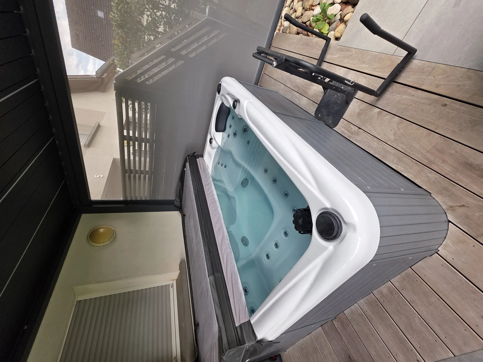 Spa gris installé sur une terrasse en bois contre une maison, protégé par une paroi vitrée, avec son releveur de couverture.