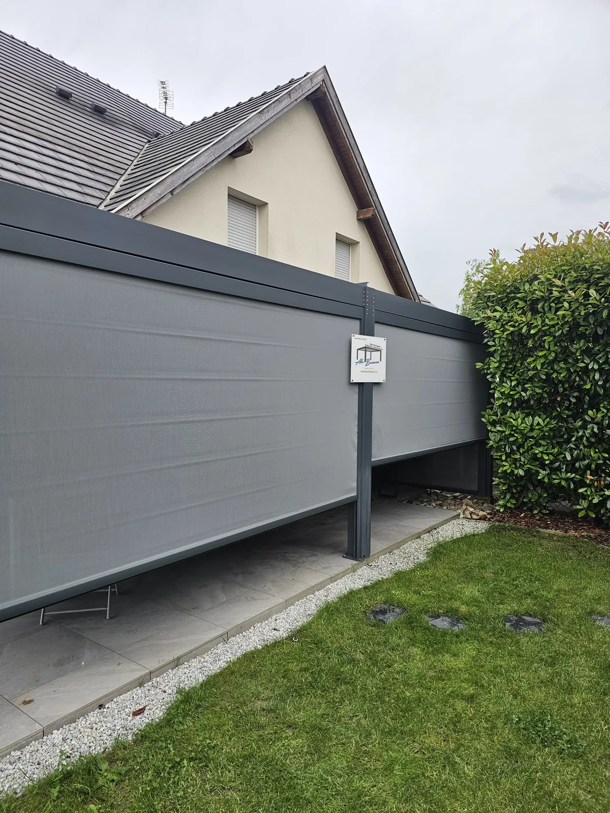 Stores zip gris baissés entre les poteaux d’une pergola aluminium anthracite, panneau Alu Business sur le poteau central, maison et pelouse autour.