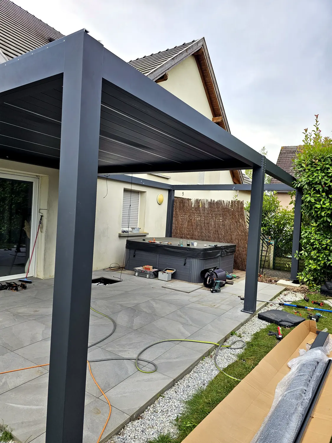 Structure aluminium anthracite lames fermées vue de dessous, poteau au premier plan, spa sous la pergola et câbles de chantier au sol.